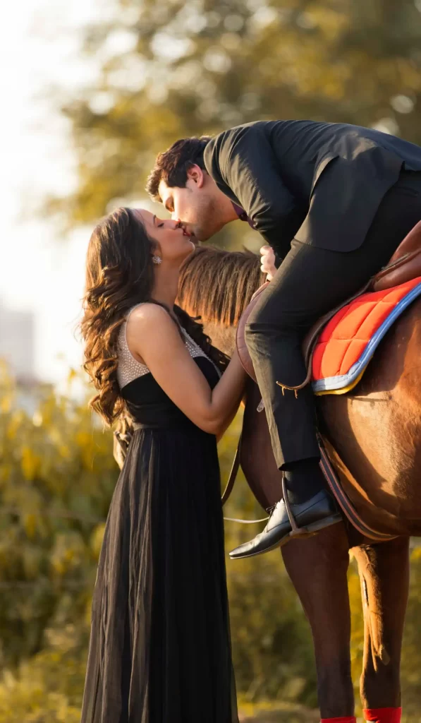 In a romantic scene, the groom sits atop a majestic horse while the bride stands nearby, sharing an affectionate kiss, capturing the timeless charm and elegance of a wedding celebration.