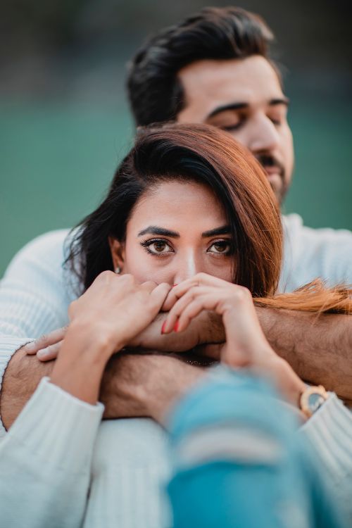 A couple standing close together, with the person wearing a suit hugging the person wearing a white dress from behind in a pre-wedding photo shoot.