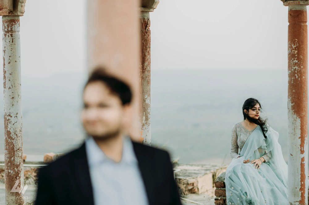 A radiant woman stands gracefully behind her partner, framed by the majestic architecture of the ancient fort, capturing a moment of timeless romance and elegance.