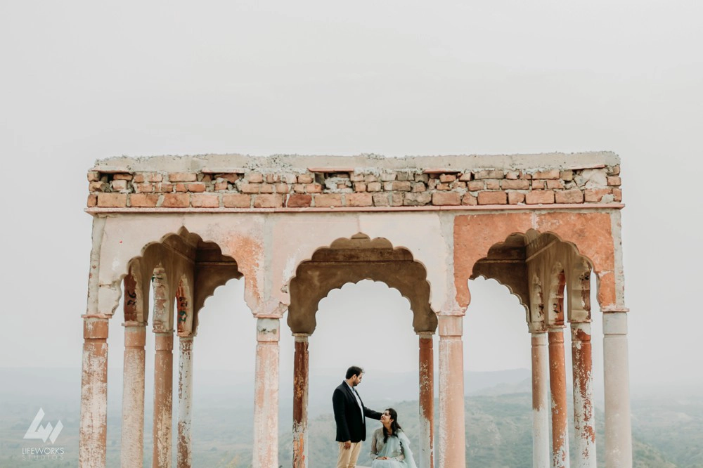 A pre-wedding couple in a picturesque setting at Tijara Fort. The woman is sitting while the man is standing in an open area surrounded by pillars.