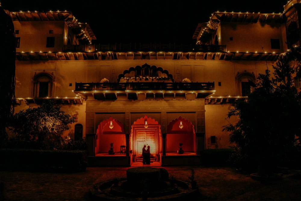 standing at majestic Tijara Fort, with the historic structure and lush greenery in the background. The couple is smiling, holding, and looking at each other.