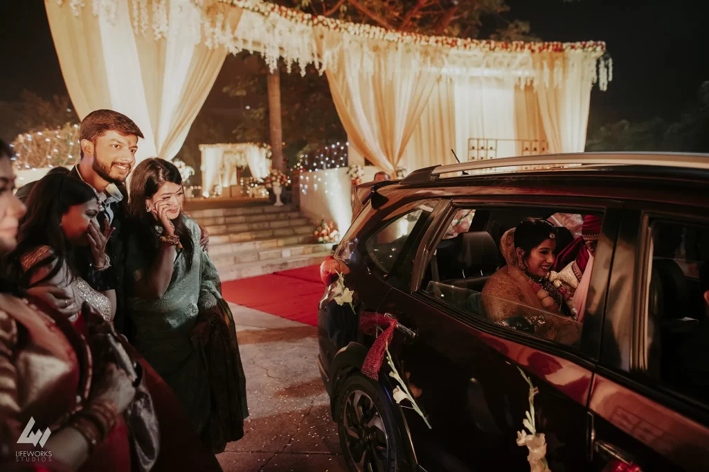 A newlywed couple dressed in traditional attire sits in a car as family members bid them a heartfelt farewell during a 'bidaai' ceremony, signifying a poignant moment in Indian wedding customs.