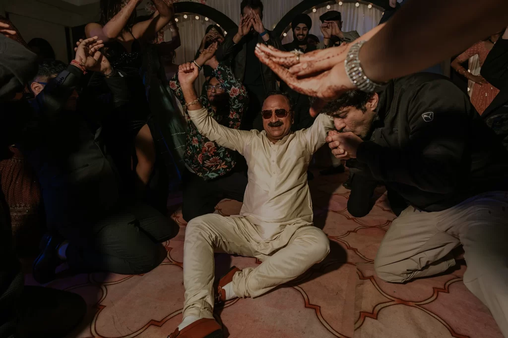  During the sangeet ceremony, the father of the groom sits on the floor, joyfully participating in the celebratory dance festivities.  