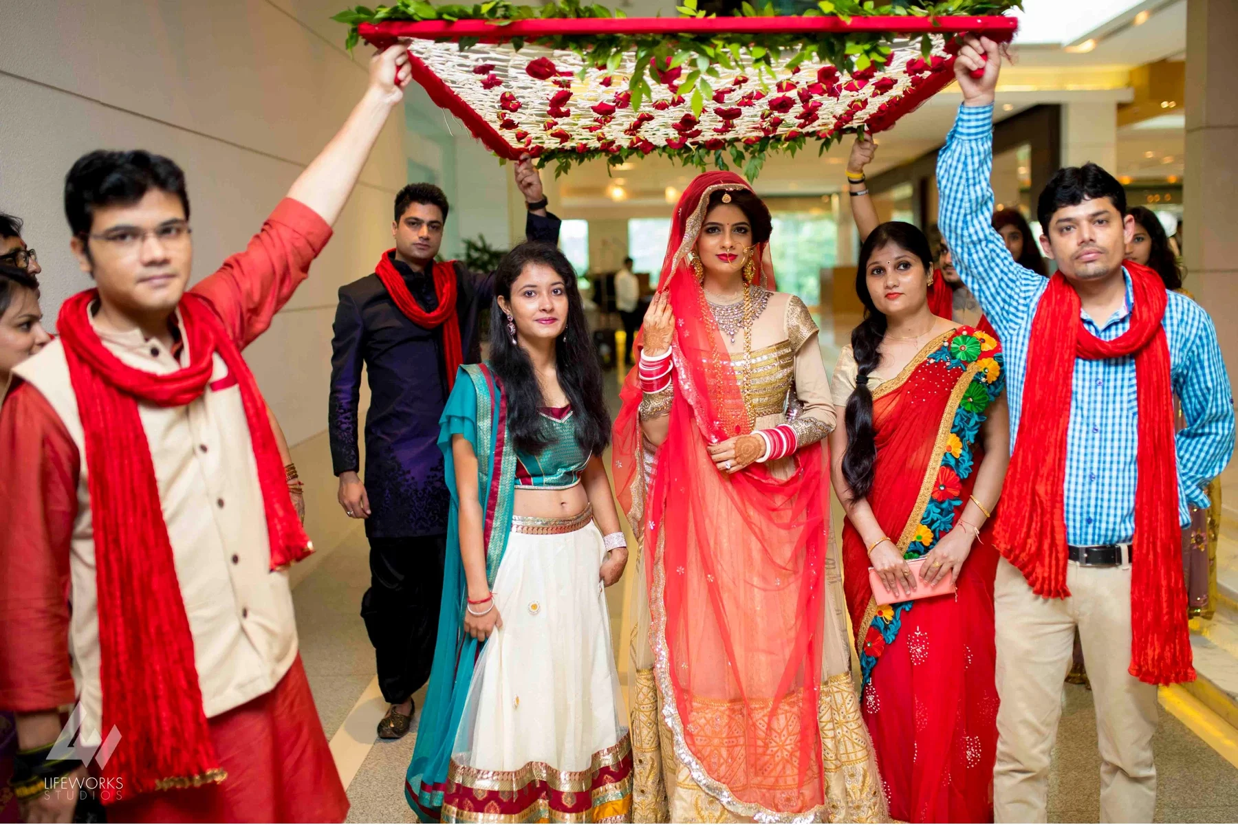 The bride in a vibrant red and gold lehenga, walking gracefully under a floral canopy during her bridal entry.