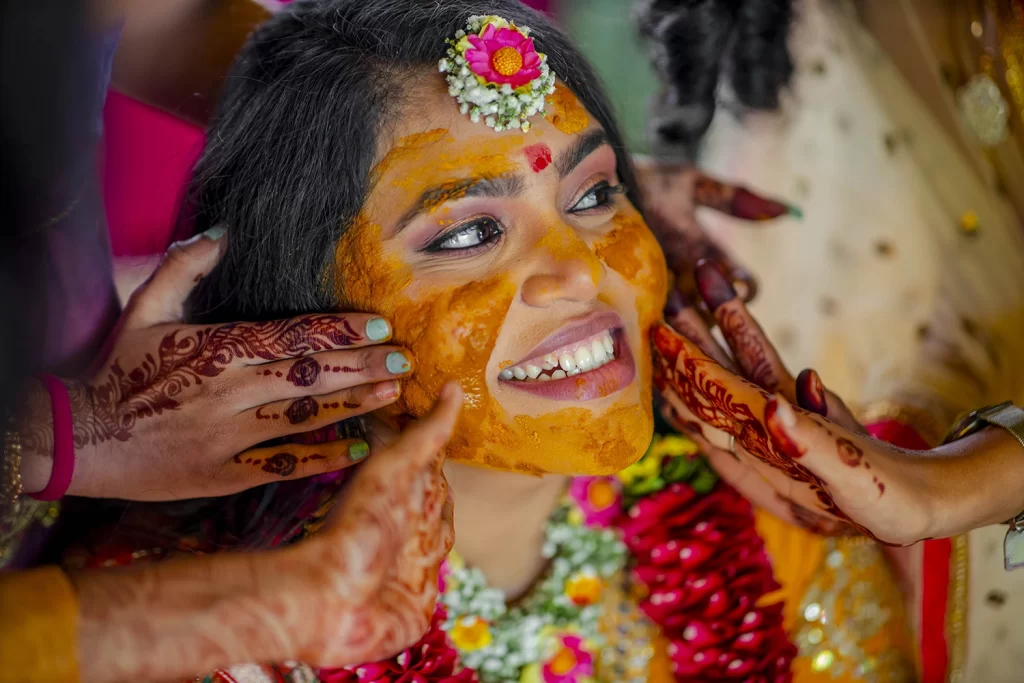 A bride smiling as her family and friends apply haldi paste on her face during the pithi ceremony