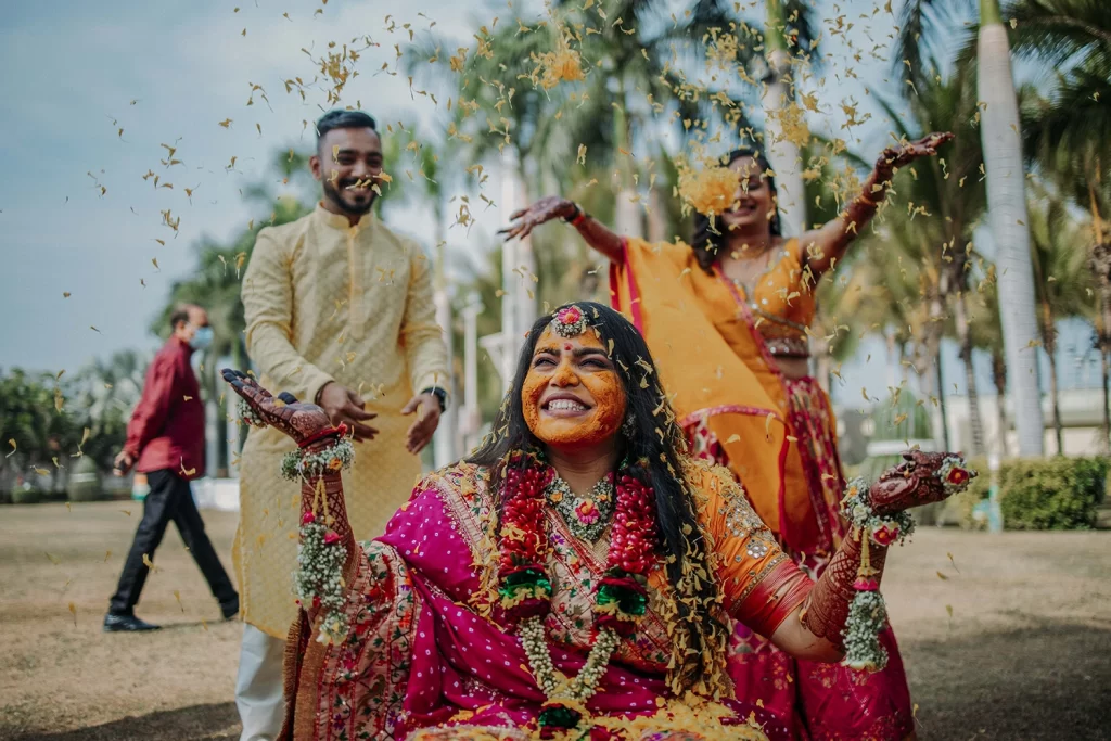 A bride-to-be sitting joyfully as friends and family shower her with vibrant flower petals during the pithi ceremony at a Gujarati wedding celebration."