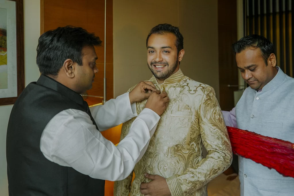 Groom getting ready surrounded by family, adjusting sherwani and looking eager, moments before his special day begins