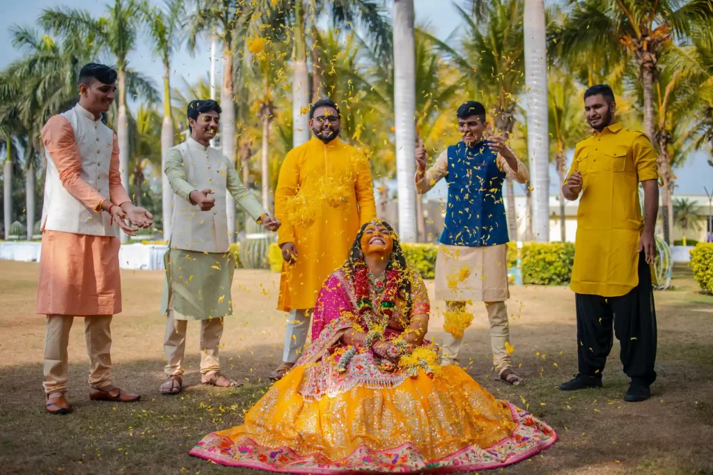 A bride-to-be sitting joyfully as friends and family shower her with vibrant flower petals during the pithi ceremony at a Gujarati wedding celebration."