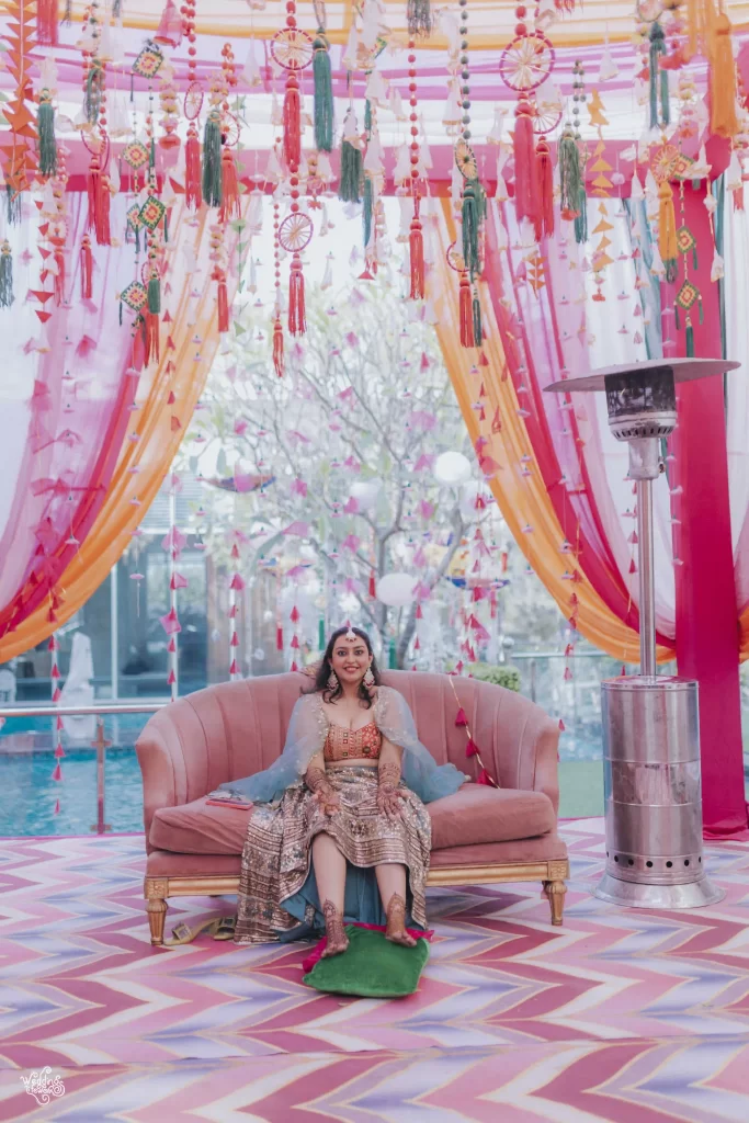 An elegant mehndi decoration featuring colorful drapes, floral arrangements, and twinkling lights at a wedding venue.